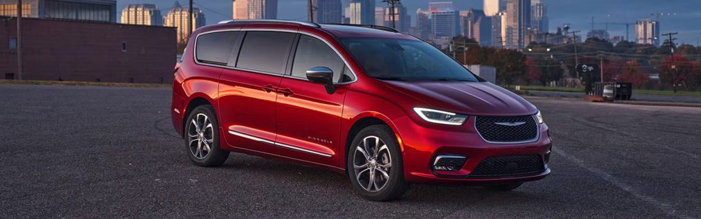 Red Chrysler Pacifica parked on a paved surface with a city skyline in the background at sunset.