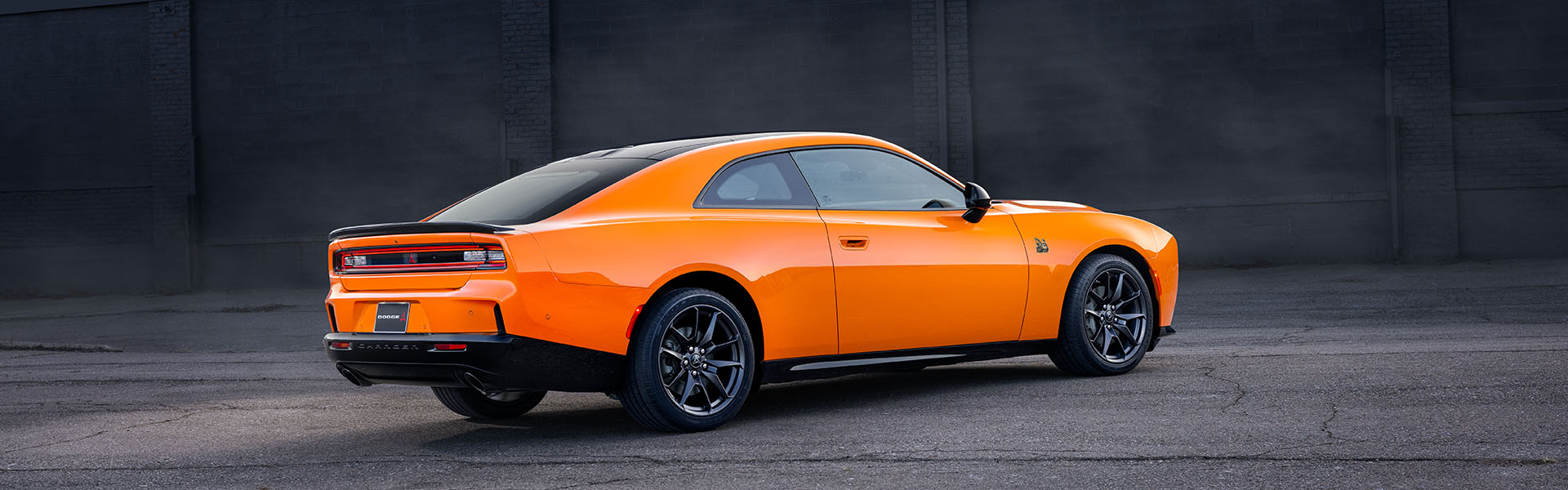 Orange 2026 Dodge Charger parked on a paved surface against a dark industrial wall backdrop.