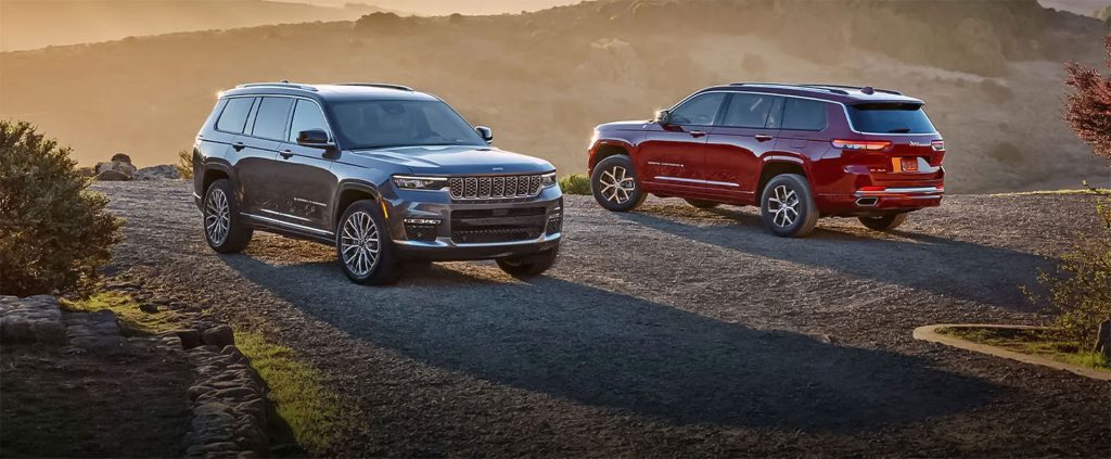 Two Jeep SUVs—one grey and one deep red—are parked on an elevated gravel trail during a golden sunset. The rolling, sunlit hills in the background create a warm, scenic atmosphere highlighting the vehicles' rugged yet luxury design.