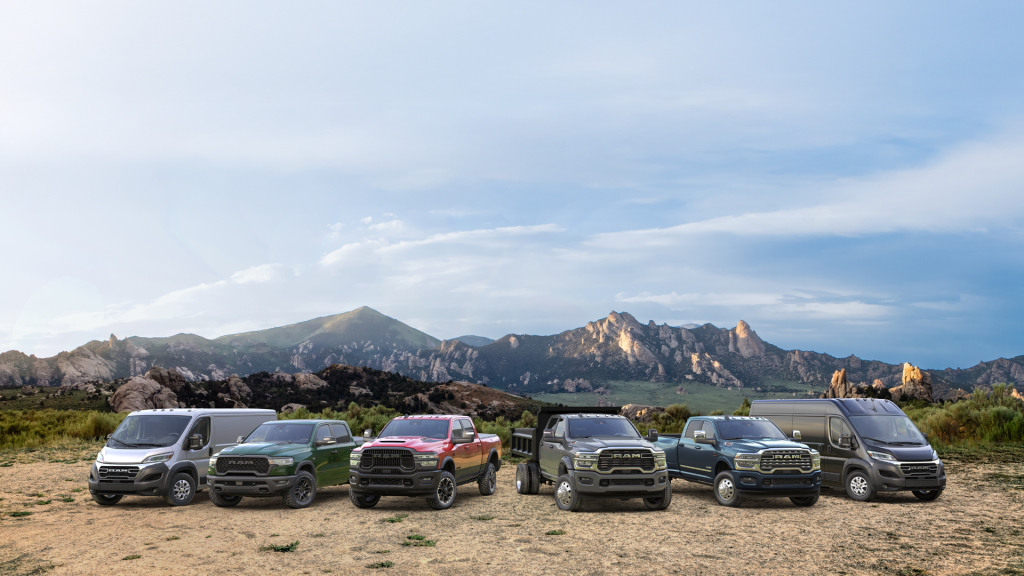 A lineup of six Ram commercial vehicles—including cargo vans, pickup trucks, and a dump truck—parked in a row on a dirt field. A dramatic mountain range stretches across the background under a bright, partly cloudy sky.