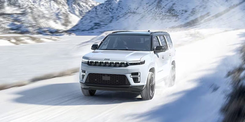 A white Jeep SUV drives through snow against a backdrop of snow-covered mountains.