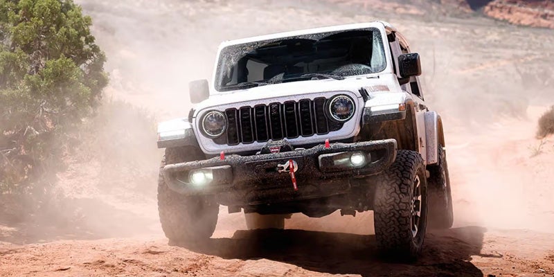A white Jeep Wrangler kicks up dust while driving across rugged terrain.
