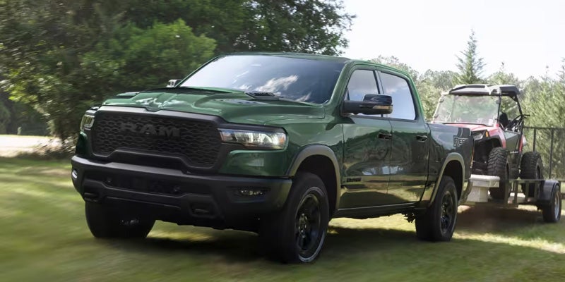 A green Ram pickup truck tows an off-road vehicle on a trailer along a tree-lined road.